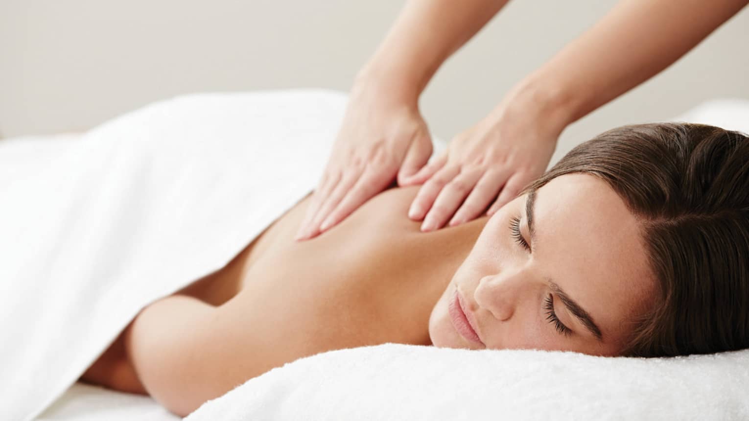 Woman with eyes closed lies under white sheet on table as hands massage her shoulders