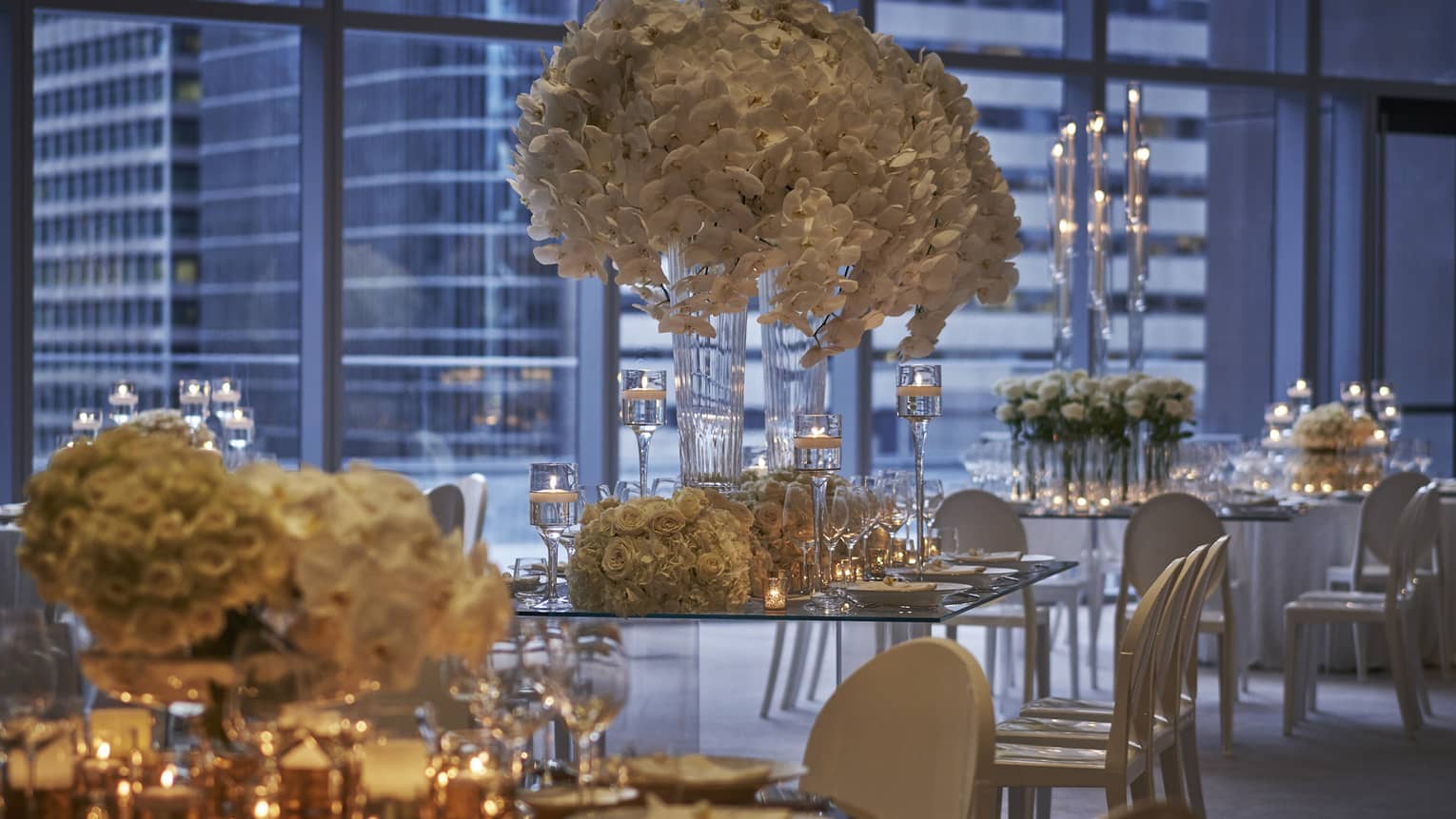 Glass tables with white chairs surrounding them, and many floral arrangements and candles placed on top.