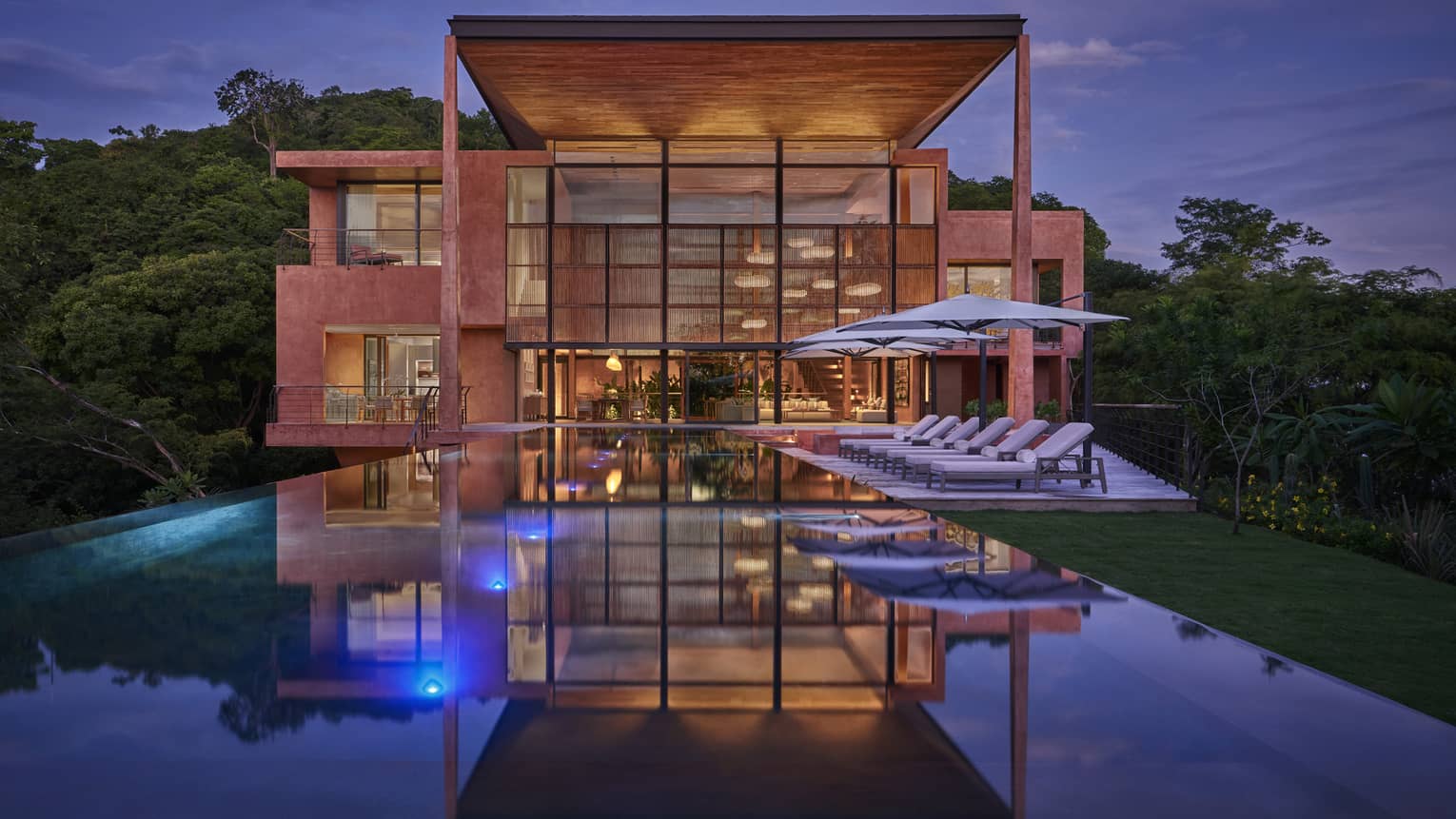 Nighttime view of the back of a private villa (lined with glass window walls); with pool and lounge chairs