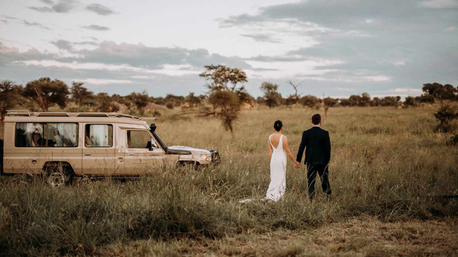 Rear view of bride and groom standing in Serengeti bushland, safari truck on left