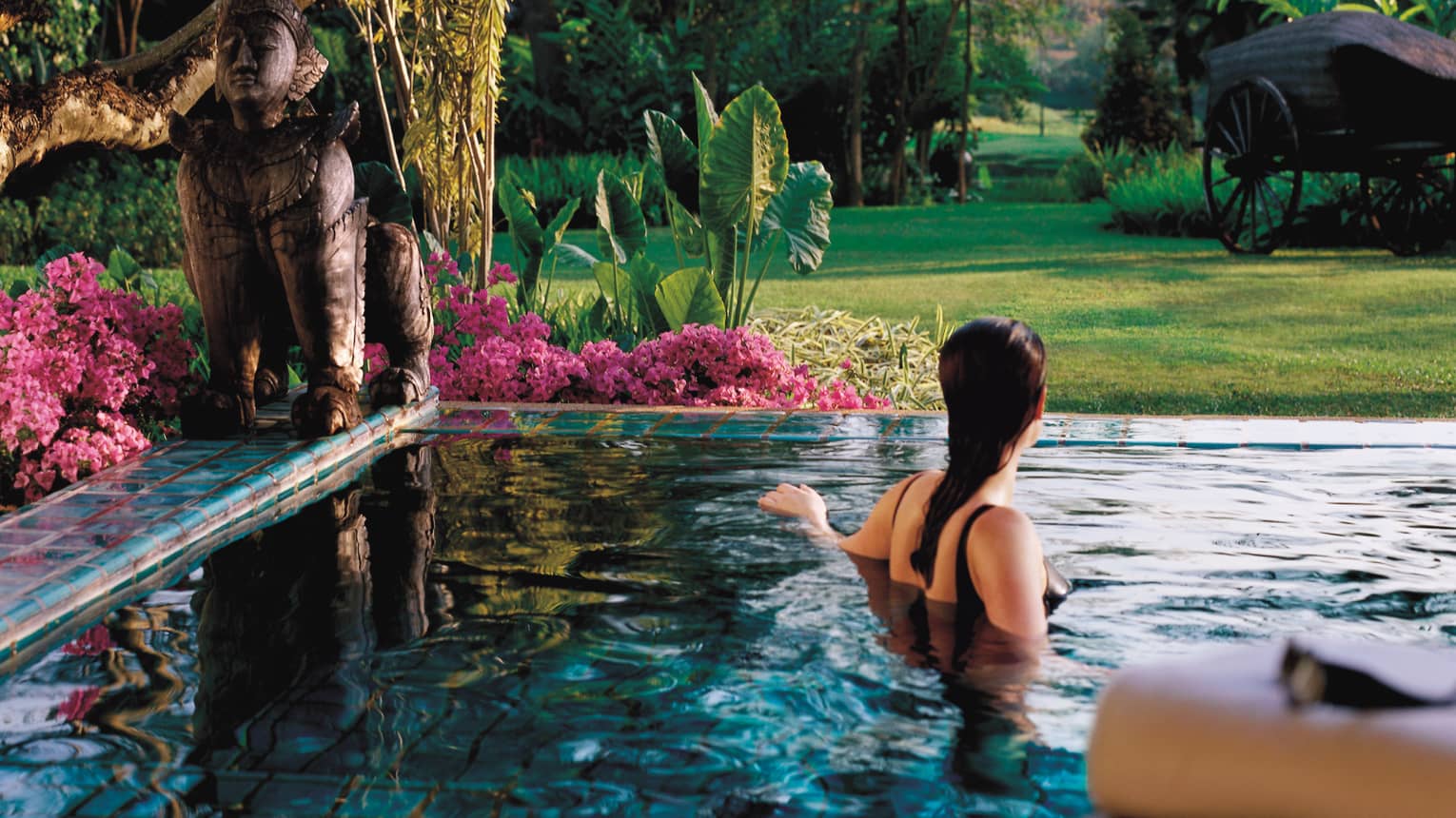 Back of woman in swimsuit wading in private plunge pool lines with pink flowers, large bronze statue