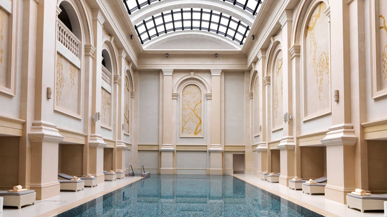 Long blue indoor swimming pool lined with deck chairs, tall cream-coloured panelled walls leading to curved sunroof 