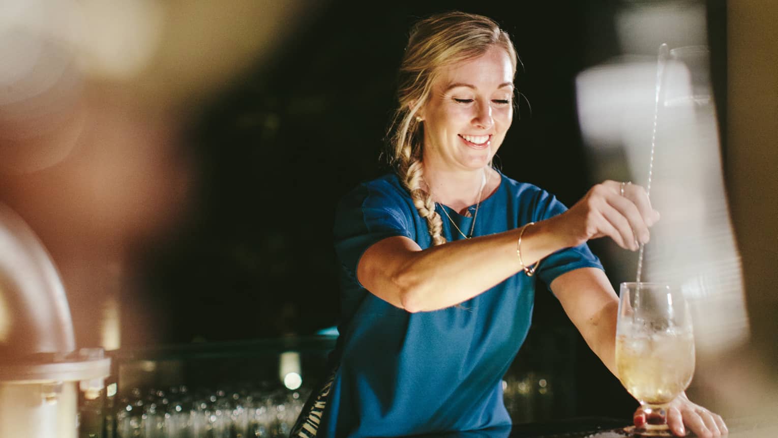 A mixologist puts together a drink at Lobby Lounge, Four Seasons Resort Maui at Wailea. She is mixing the drink and smiling.