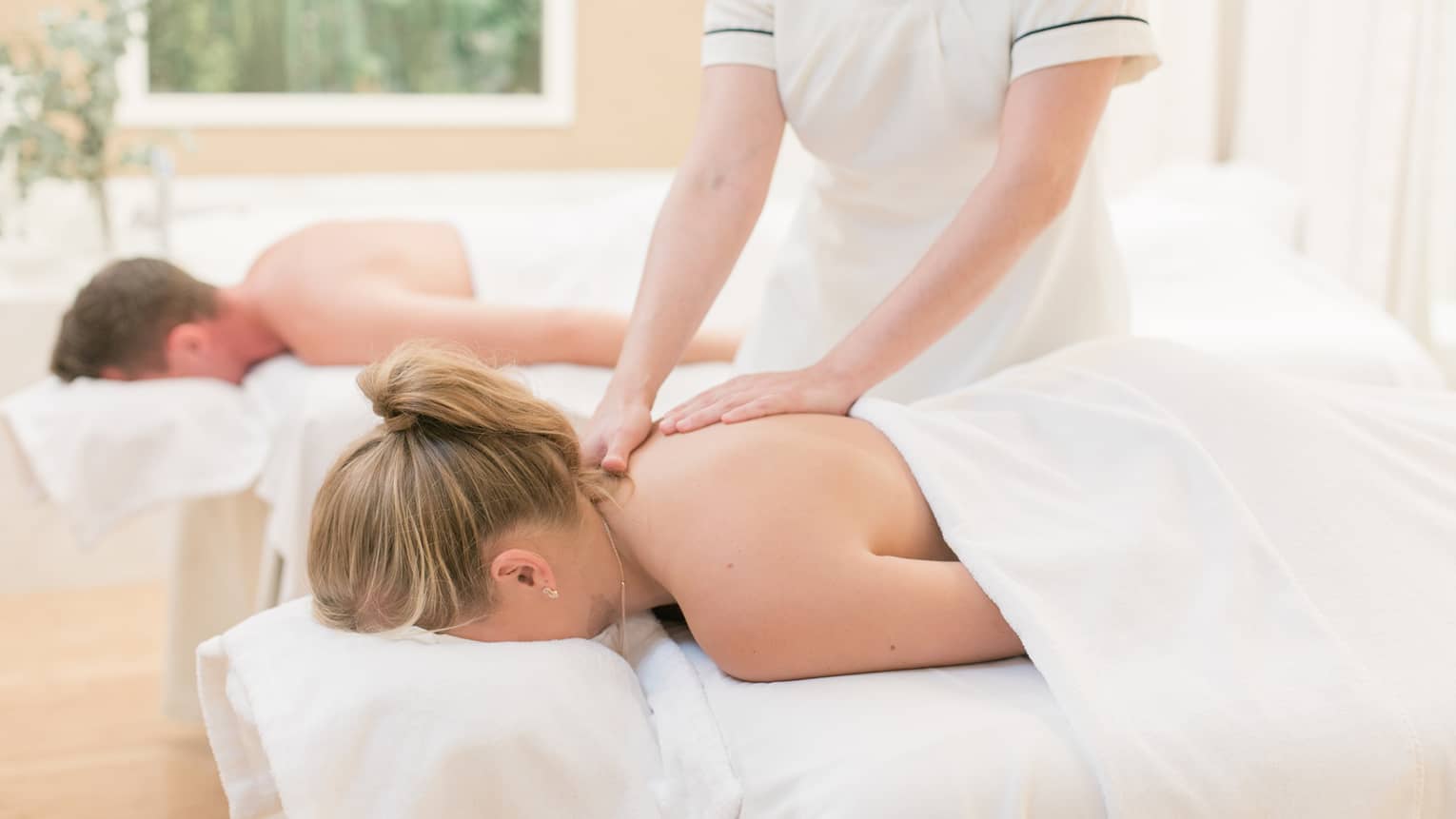 Couple laying on massage tables, spa staff massages woman's shoulders 