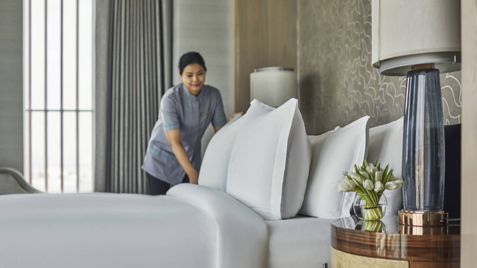 A room service attendant fluffing pillows on a freshly made bed
