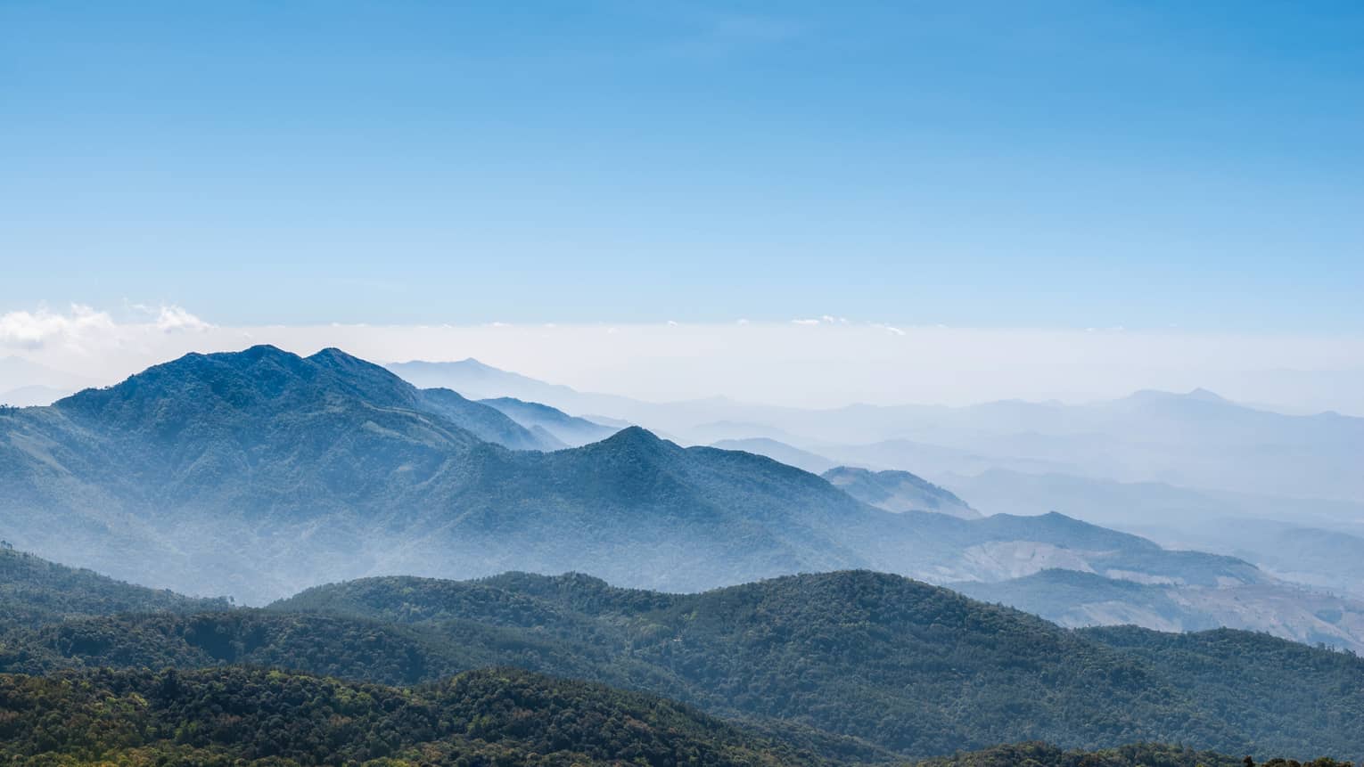 Close-up of smooth, forested mountains receding into the distance under clear sky and a misty light blue haze.