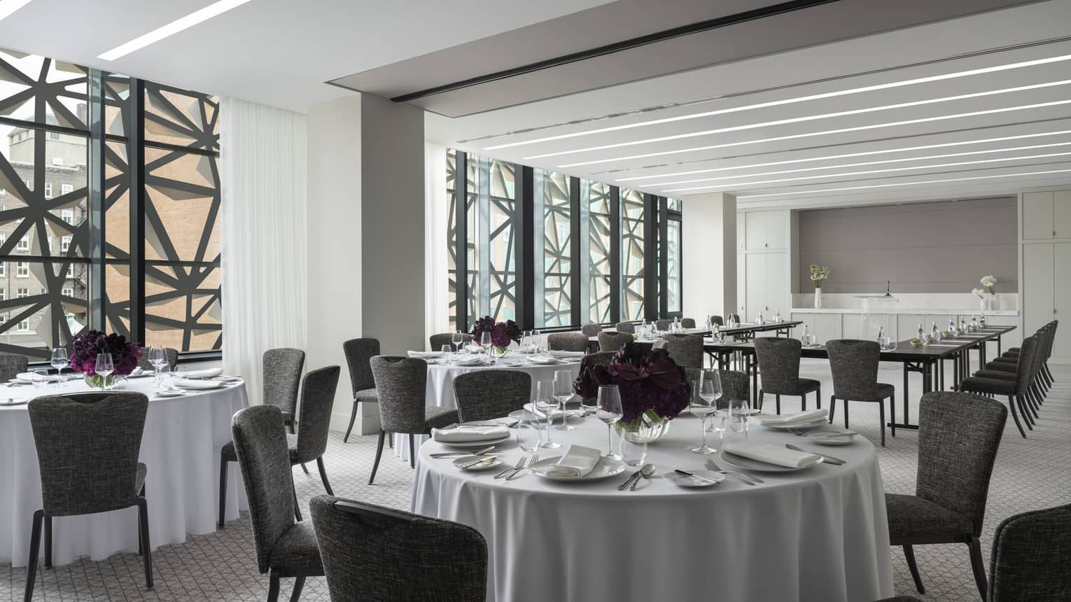 Salon with white round tables and flowers, grey chairs, U shaped table in background, ceiling-to-wall windows