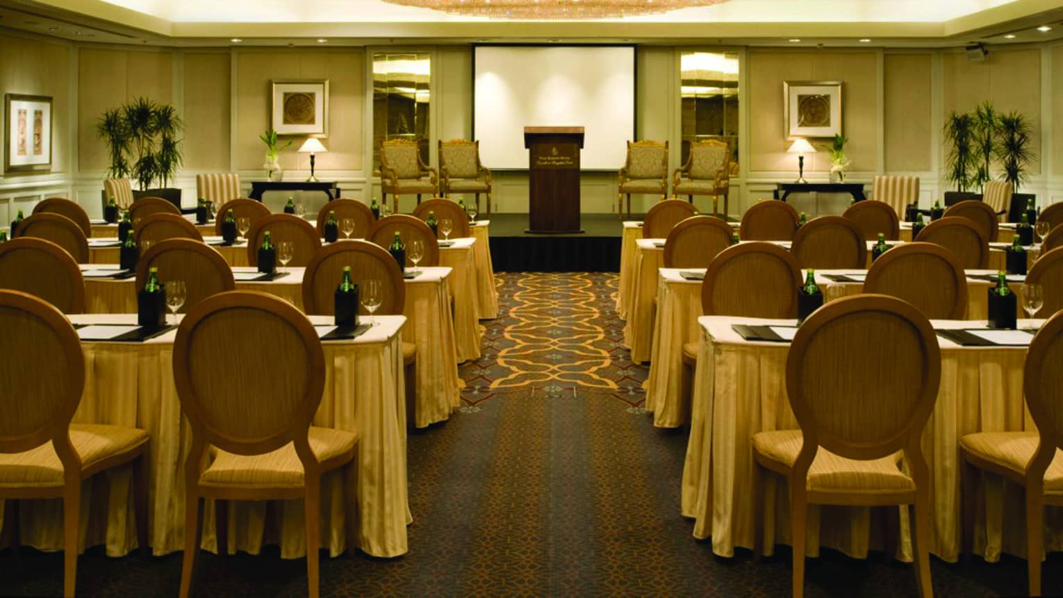 Meeting room with large chandelier, rows of tables with chairs facing podium with four chairs