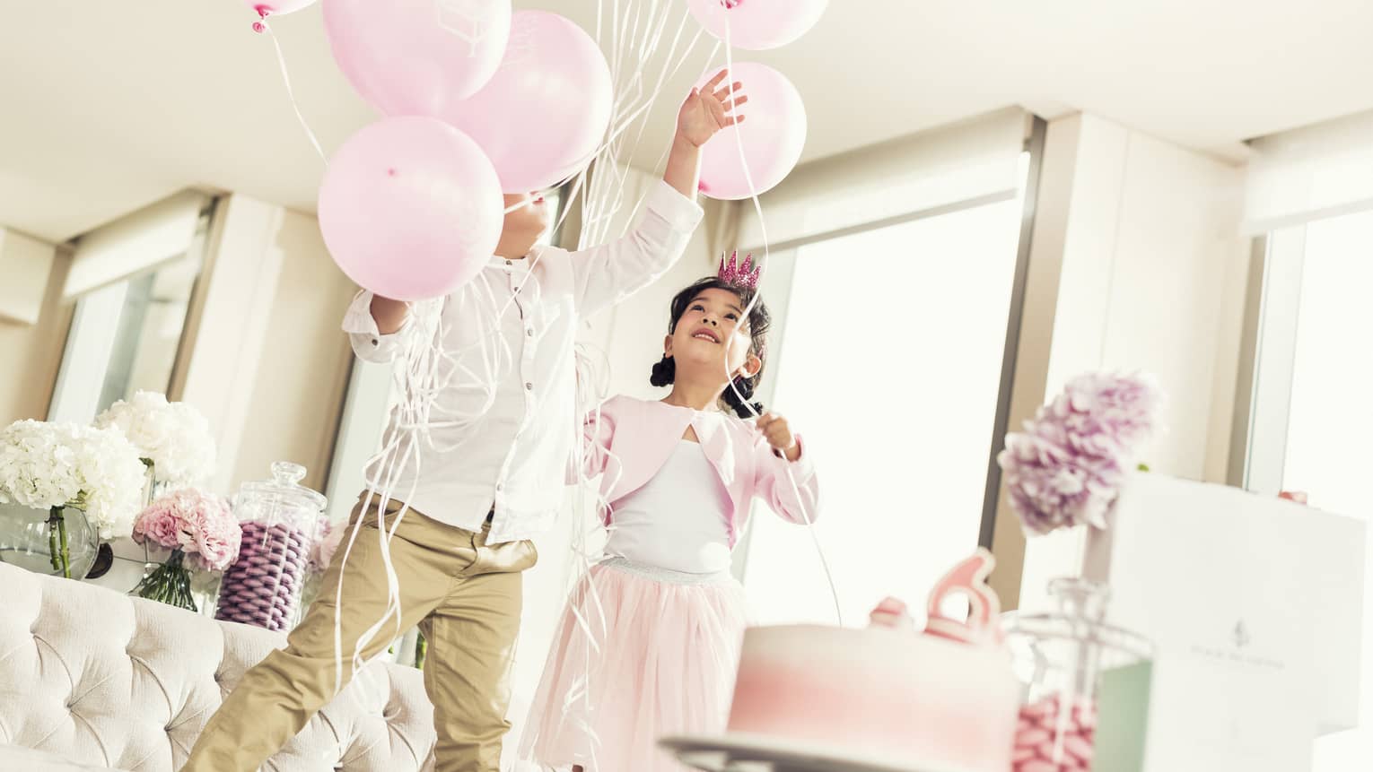 Two kids playing with pink balloons near a pink cake.