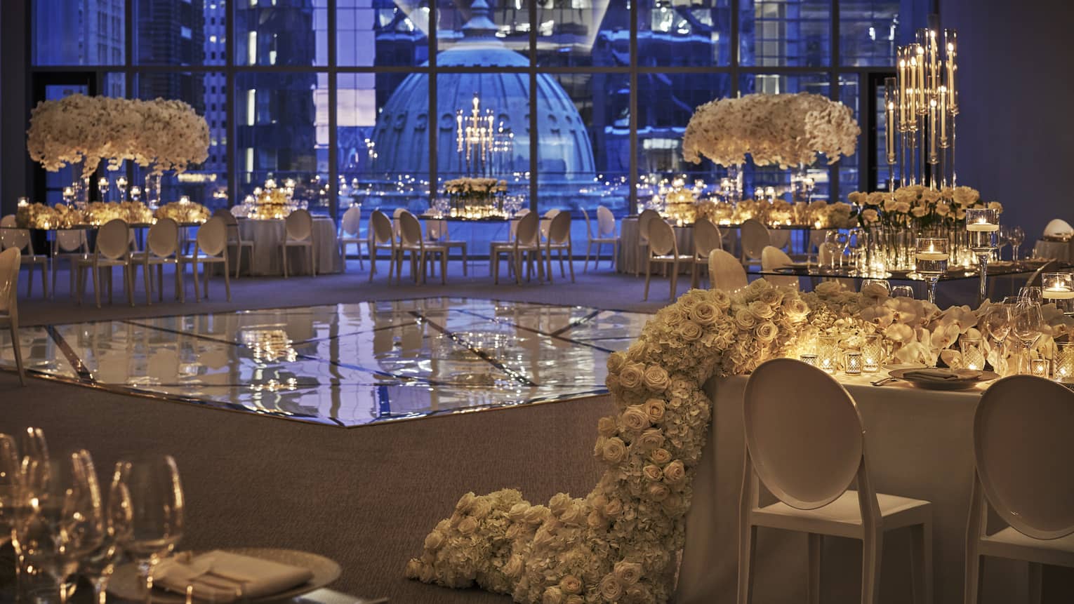 The dance floor in the ballroom is surrounded by white tables decorated with extravagant white flowers and set with white and clear dishes and glasses and overlooks downtown philadelphia through floor to ceiling windows