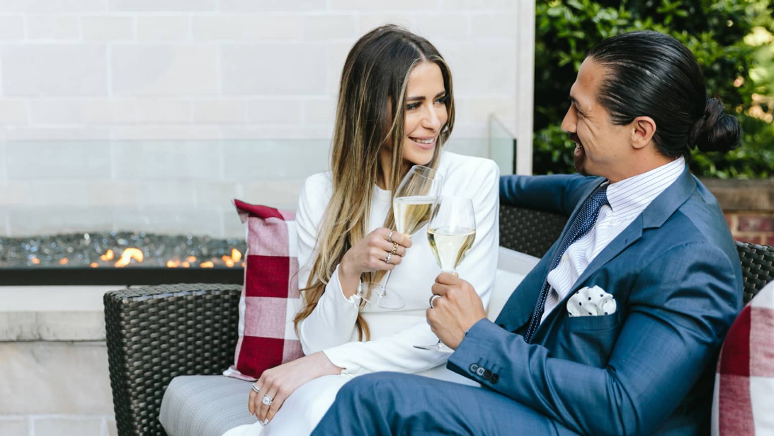 A woman in a white suit and a man in a gray suit look at each other smiling as they sip white wine on an outdoor couch during daylight.