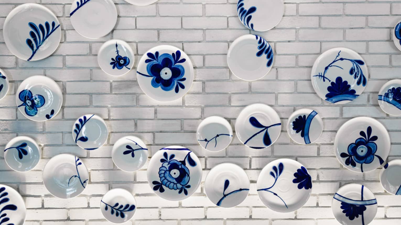 Wall art made up of various white plates painted with blue leaves and flowers hang on a white brick wall