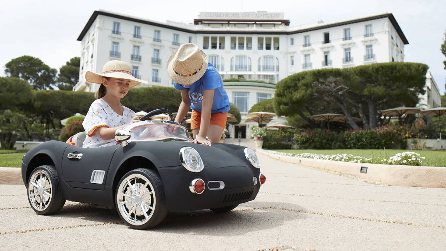 Girl wearing floral dress and straw hat driving black luxury mini convertible, boy in straw hat leaning over, hotel in backdrop