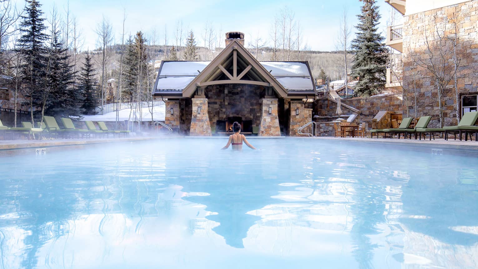 A woman in a pool with a cabin in front of her and snow covered trees around her.