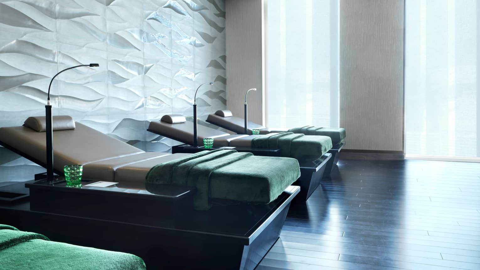 Four spa beds side-by-side with reading lamps, green blankets and glasses, in front of white wall and sunny windows