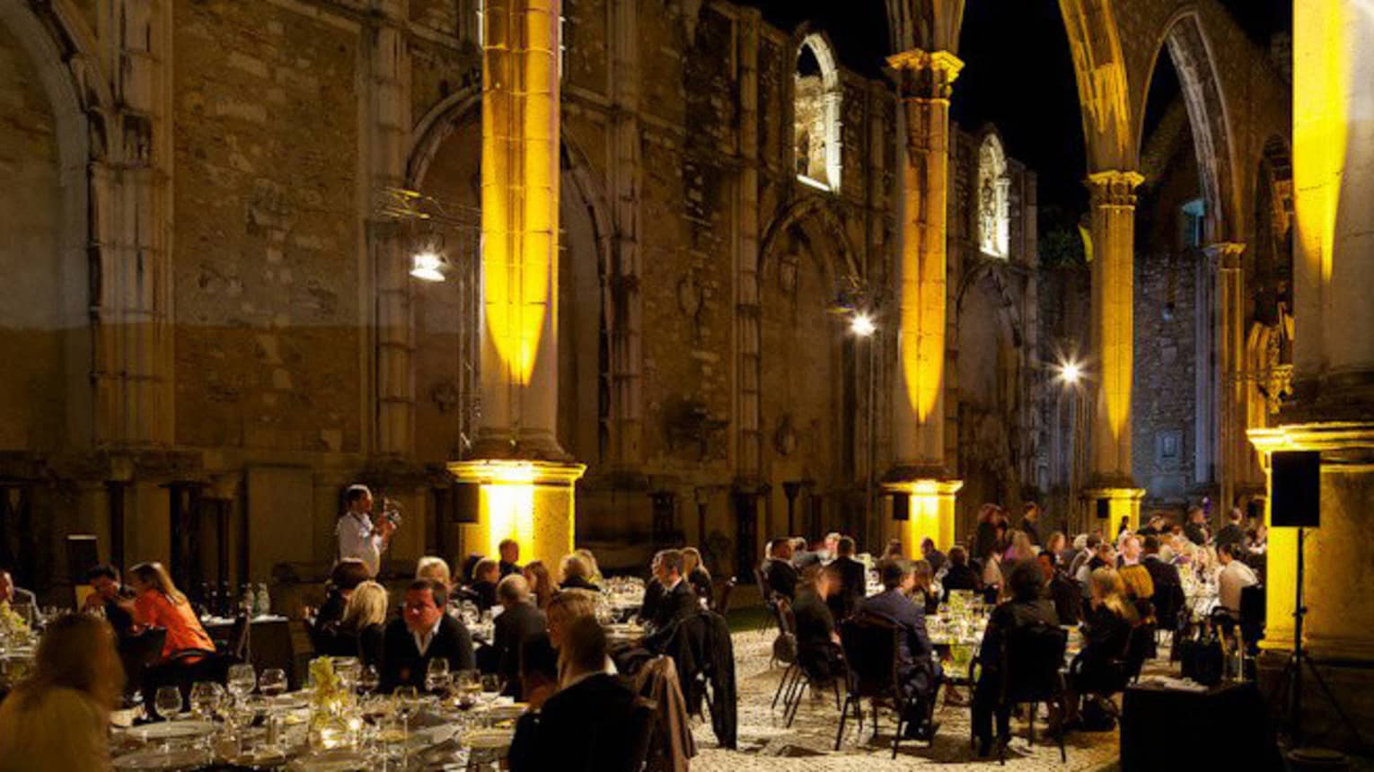 Guests dine at round banquet tables under large historic stone building at night