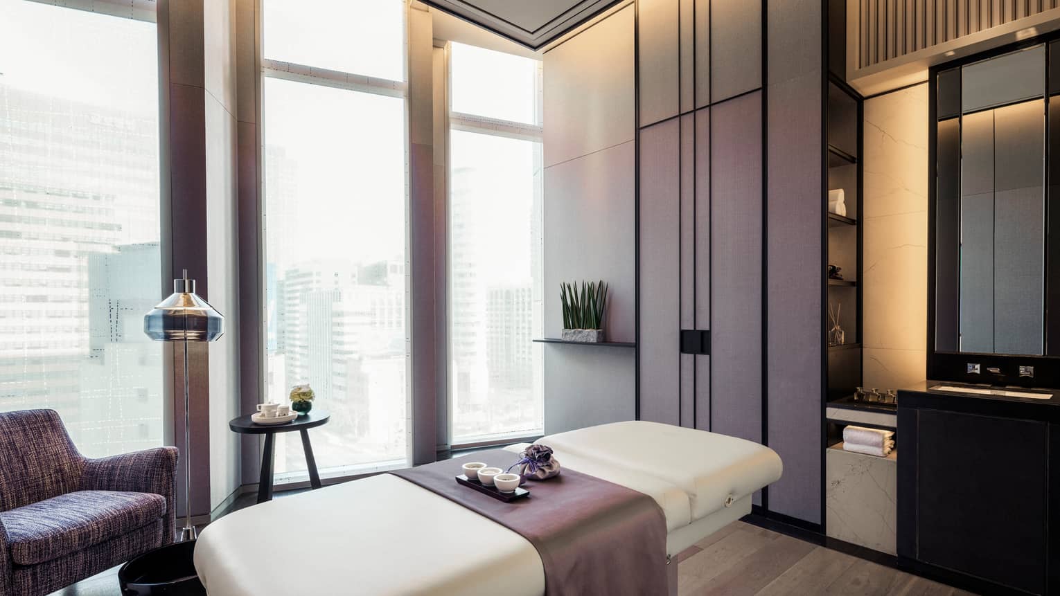 Purple accent chair, blanket over massage table with Spa tray by floor-to-ceiling windows, shades