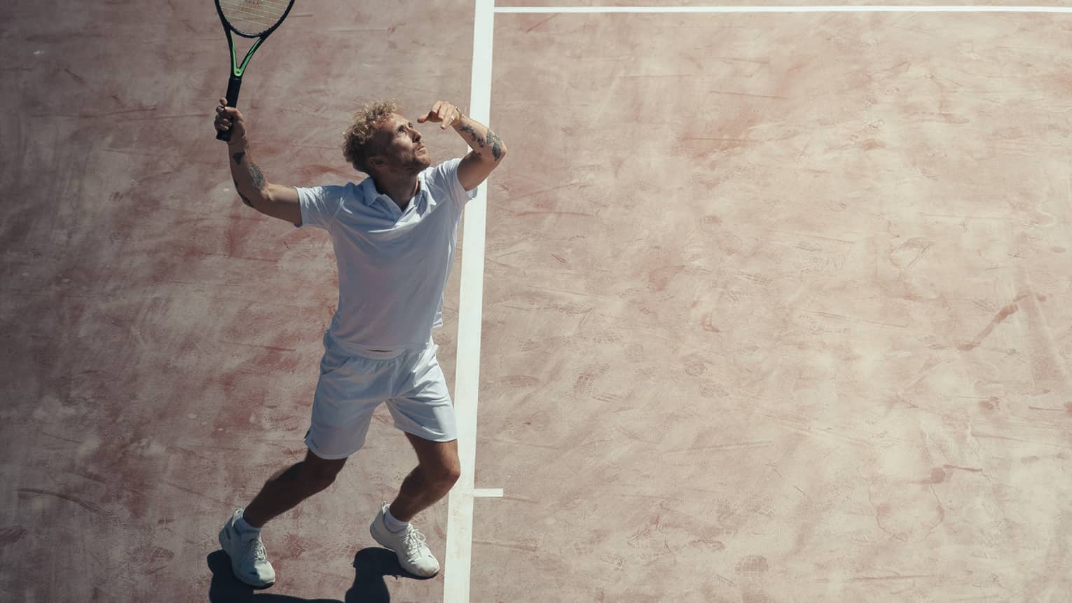 Man in white outfit playing tennis on reddish clay court