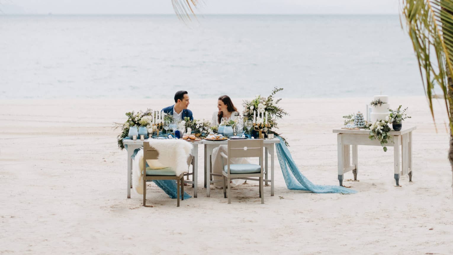 Smiling couple at private dining on white sand beach past palm leaves
