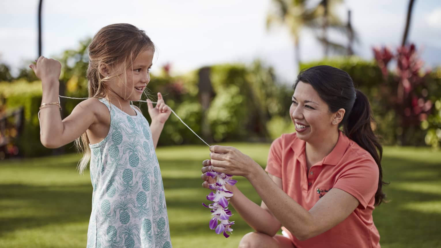 Girl in blue patterned dress smiling with staff member in orange polo, making lei