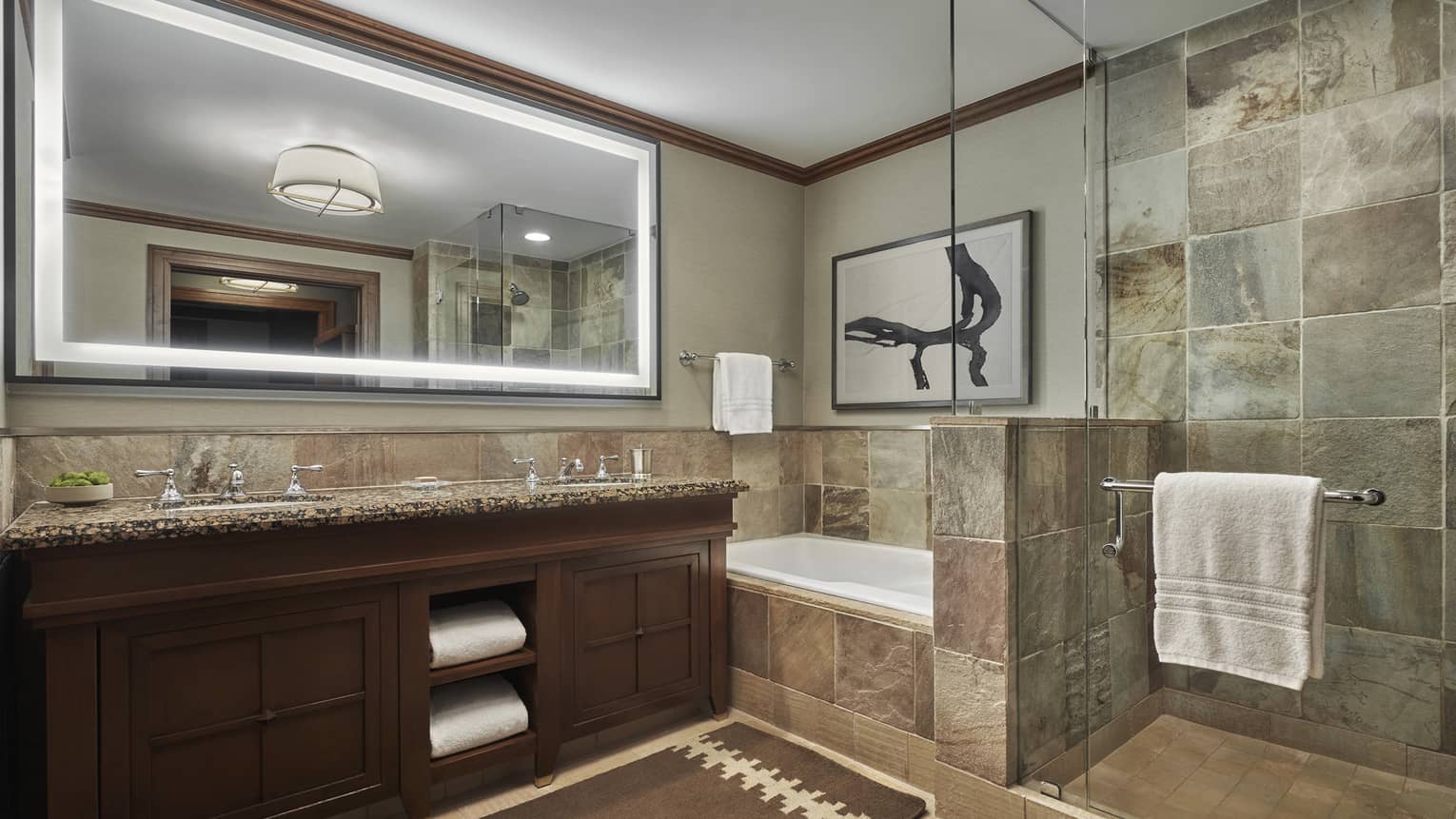 A tiled bathroom, with a double vanity, large lighted mirror and a shower and tub separated by a glass partition