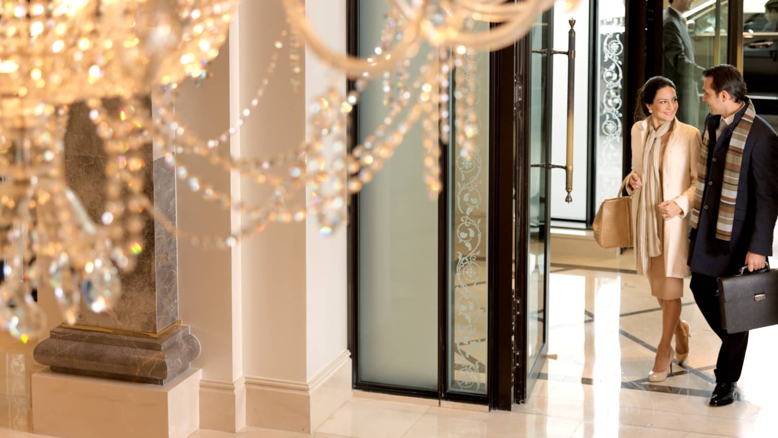 Close-up of crystal chandelier, view below of elegantly-dressed woman linking arms with man, walking in through hotel lobby doors