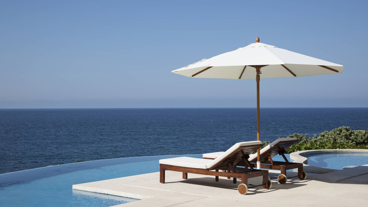 Two lounge chairs under white patio umbrella at edge of infinity pool, ocean views
