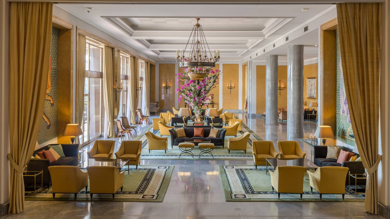 Bright hotel lobby with long gold curtains and armchairs, seating area under chandelier
