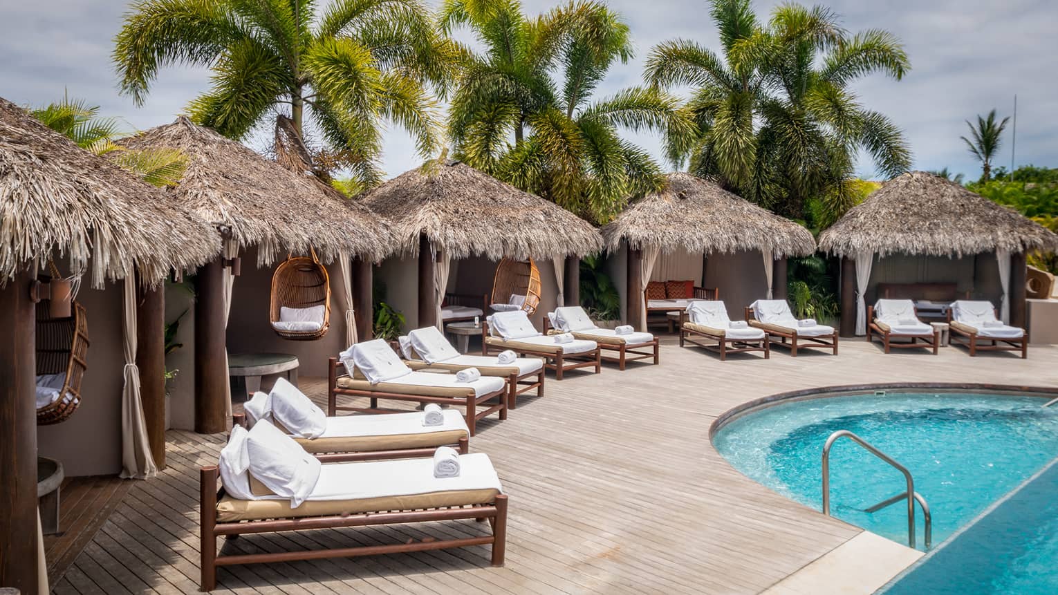 Grass-topped poolside cabanas with row of lounge chairs in front, palm trees in distance