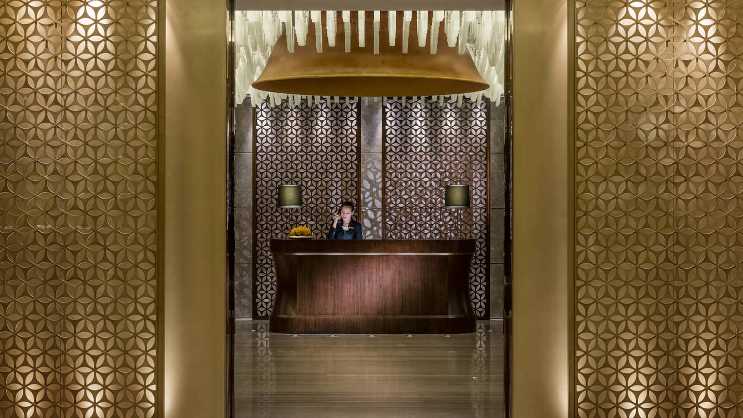 Decorative gold screens to hall where woman answers phone at wood reception desk with lamps