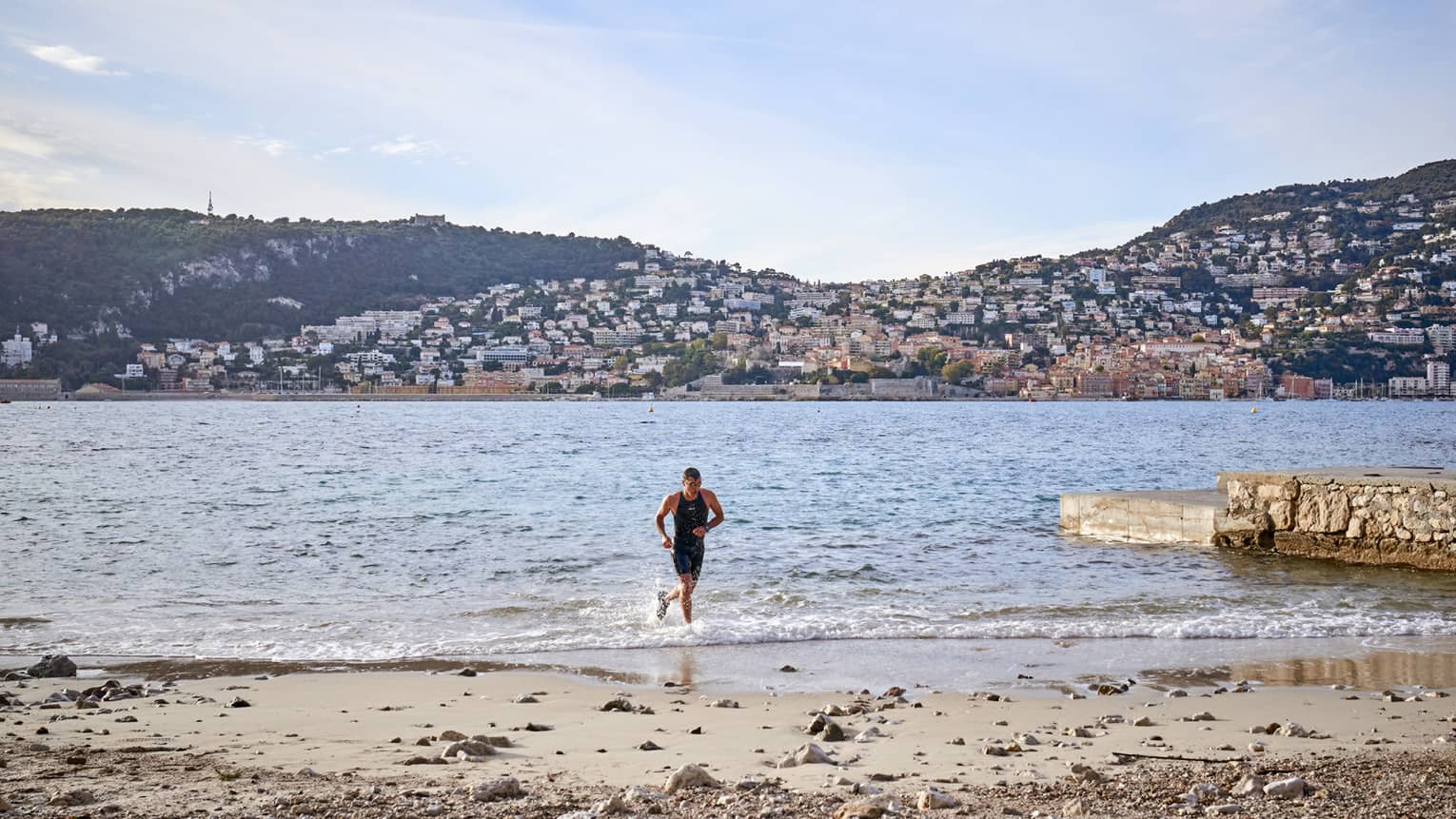 Man jogging in water toward sandy shore, hills in backdrop