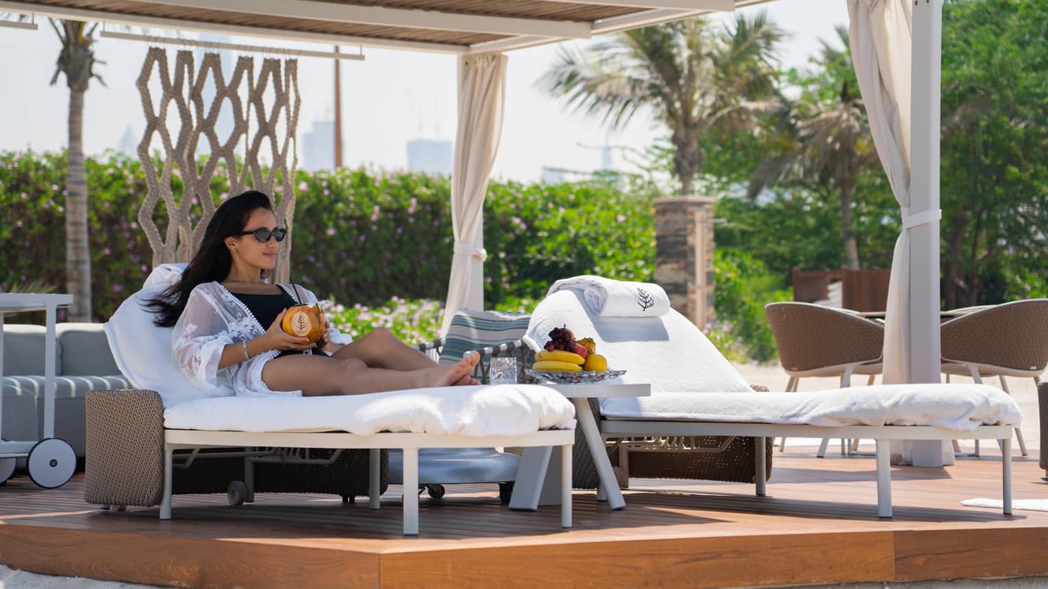 Woman in sunglasses and bathing suit lounges on chaise under poolside private cabana