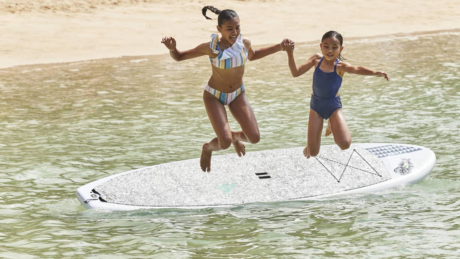 Two young girls jump hand-in-hand into the water from a stand-up paddleboard