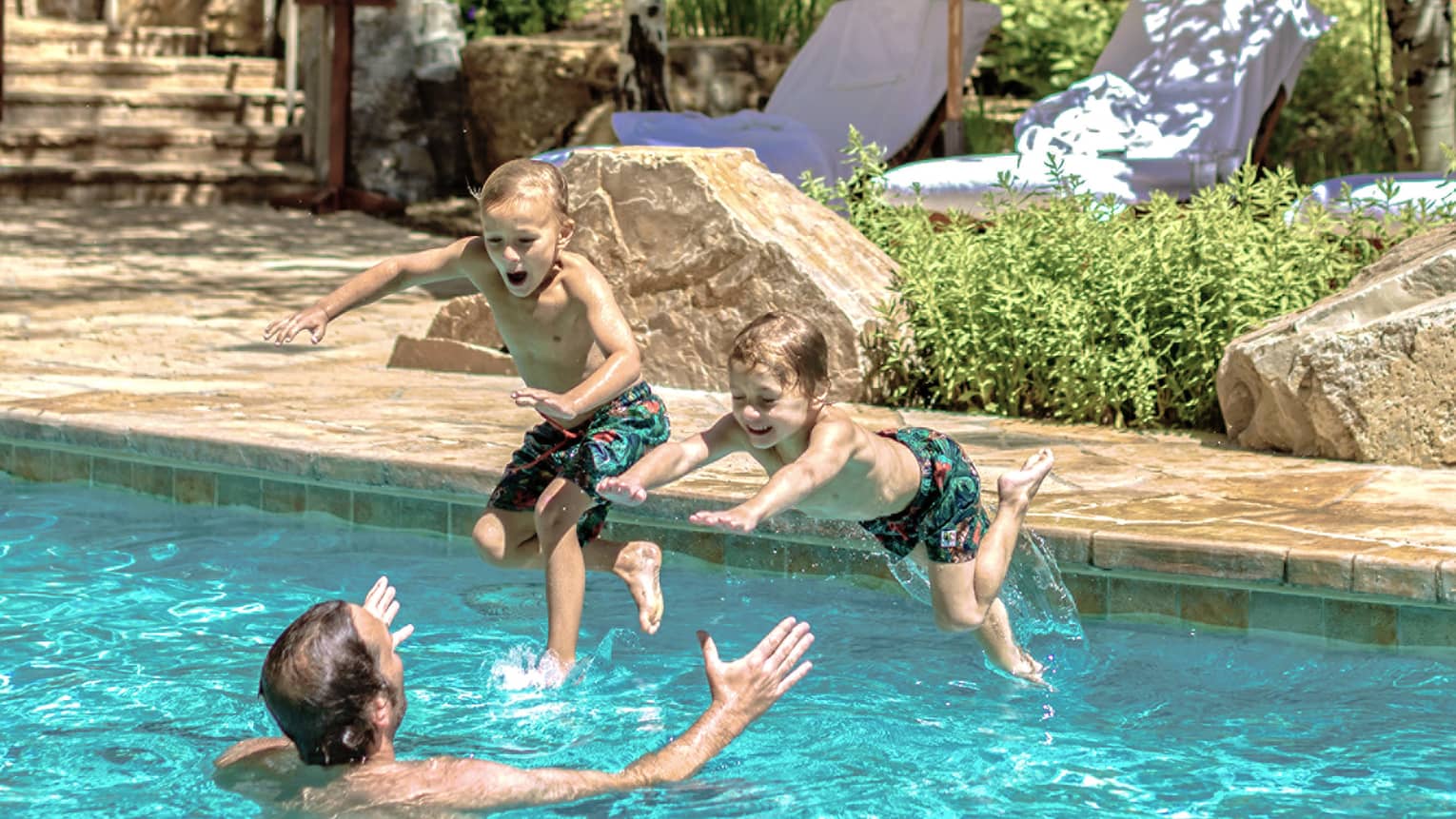 Two young boys jump into their father's outstretched arms in an outdoor pool 