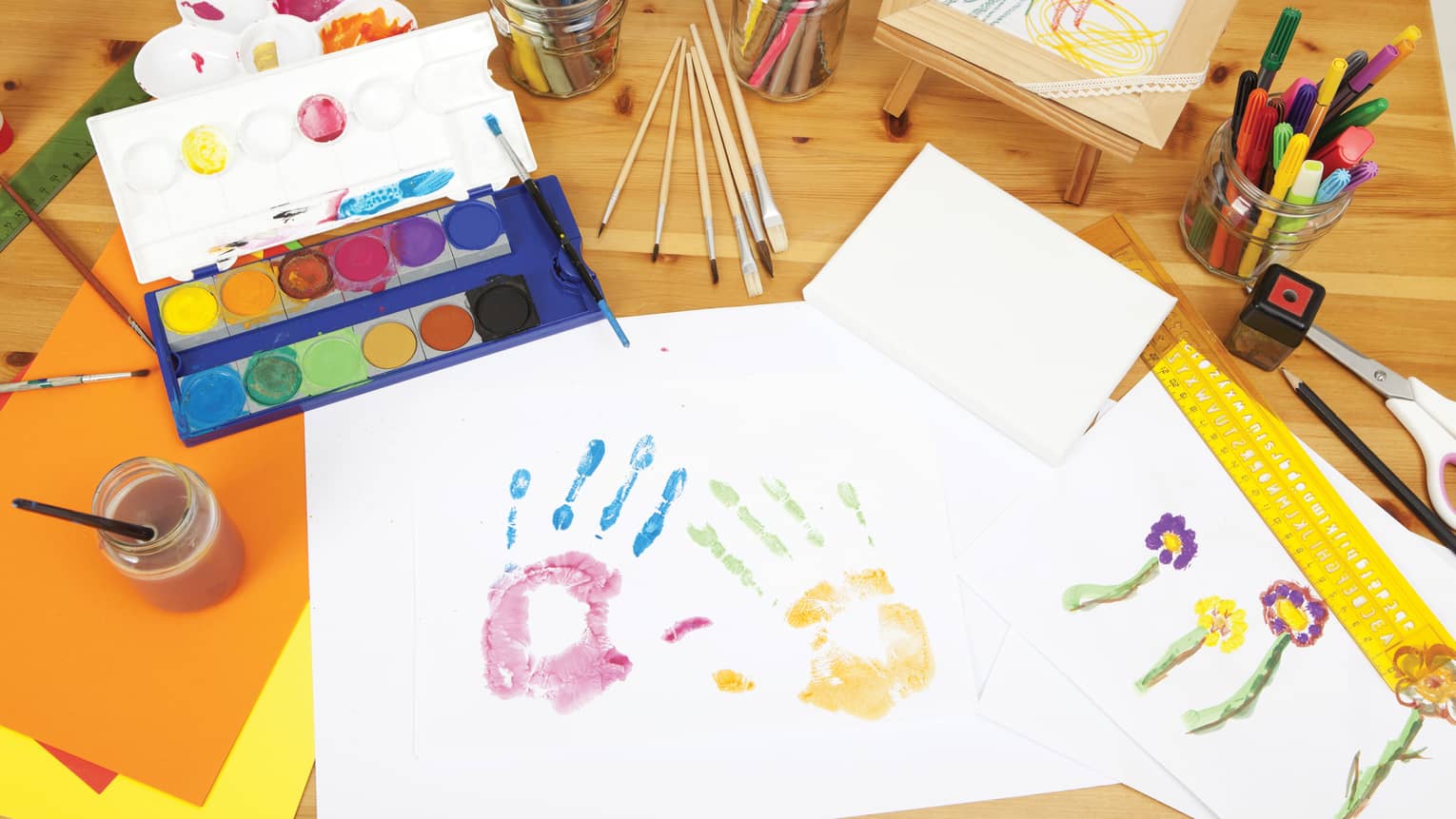 Two colourful paint handprints on paper next to tray of kid's paints on Kids Club craft table