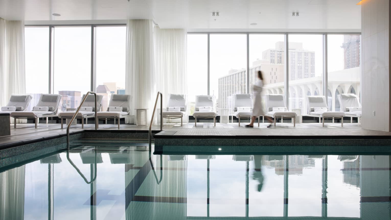 A woman in a spa robe walking along lounge chairs by an indoor pool.