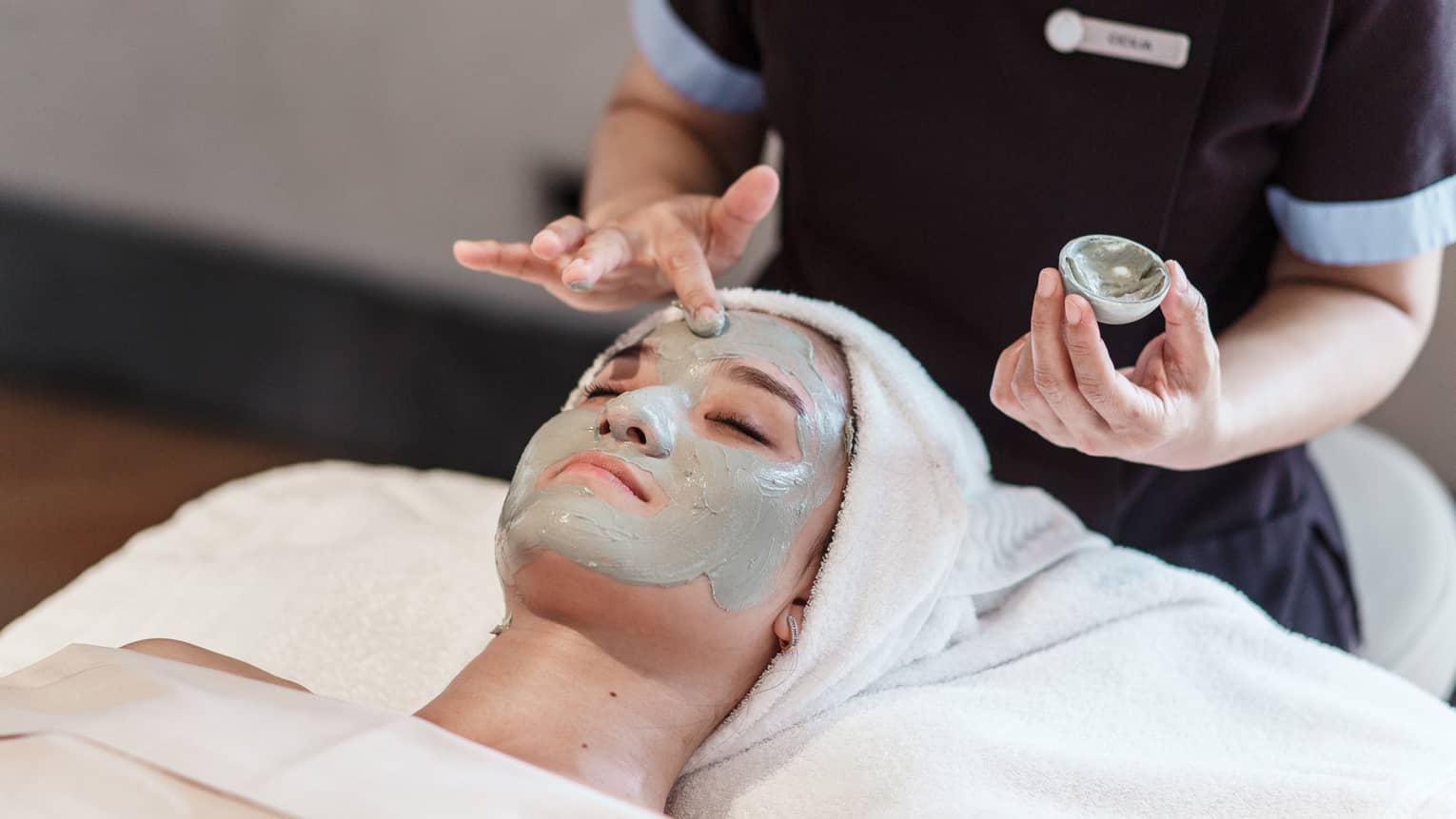 Spa staff applies green clay mask to woman's face as she lays on treatment bed 