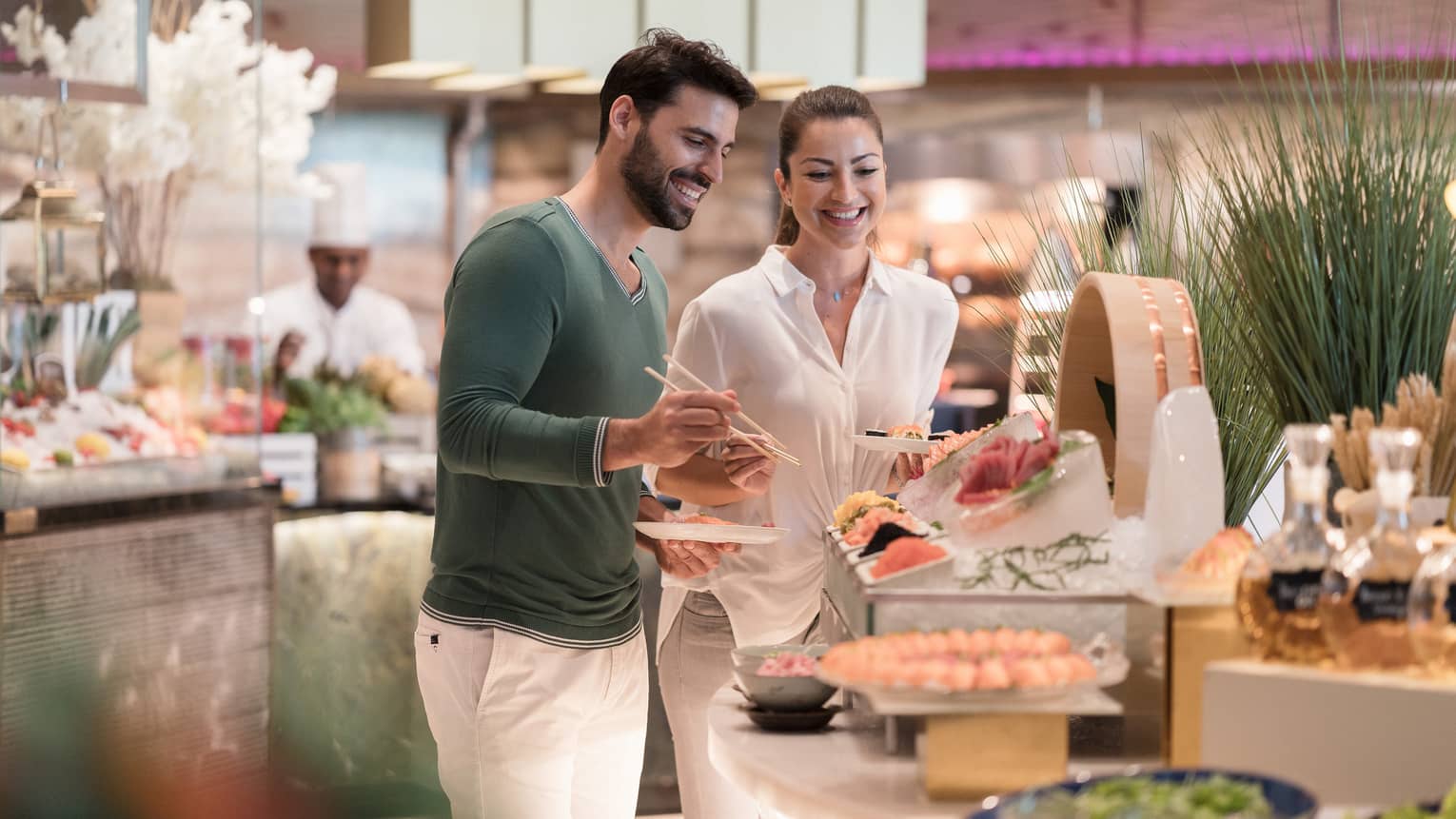 A man wearing white pants and green sweater stands next to a woman wearing all white at the sushi table of a buffet