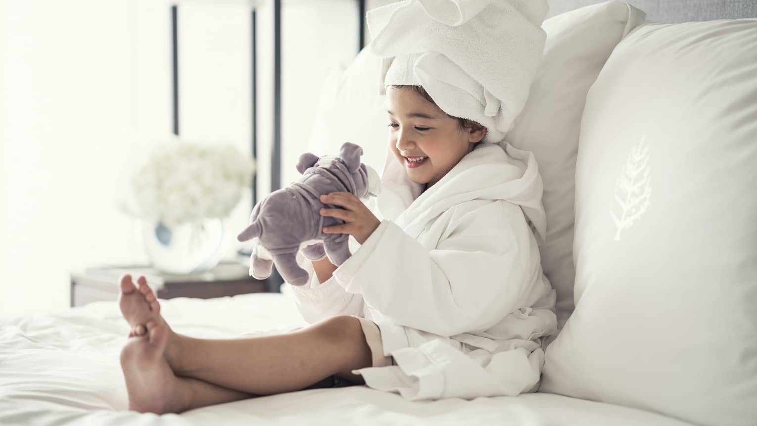 Young girl wearing a white bathrobe with towel wrap plays with plush dog toy on bed