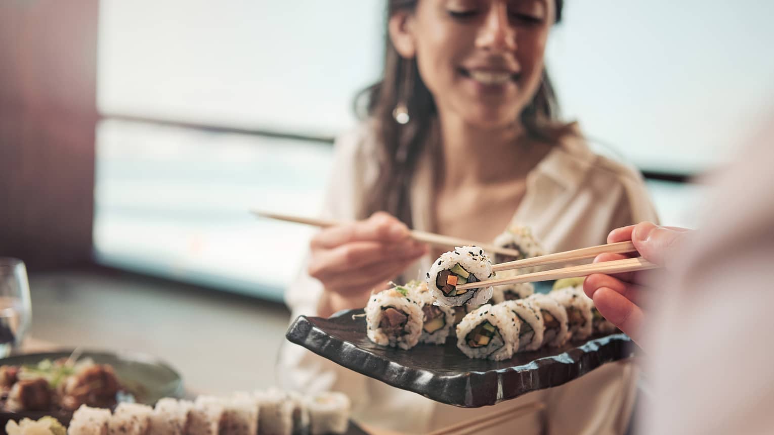 A woman picks up a sushi roll with a set of chopsticks off of a tray of sushi.