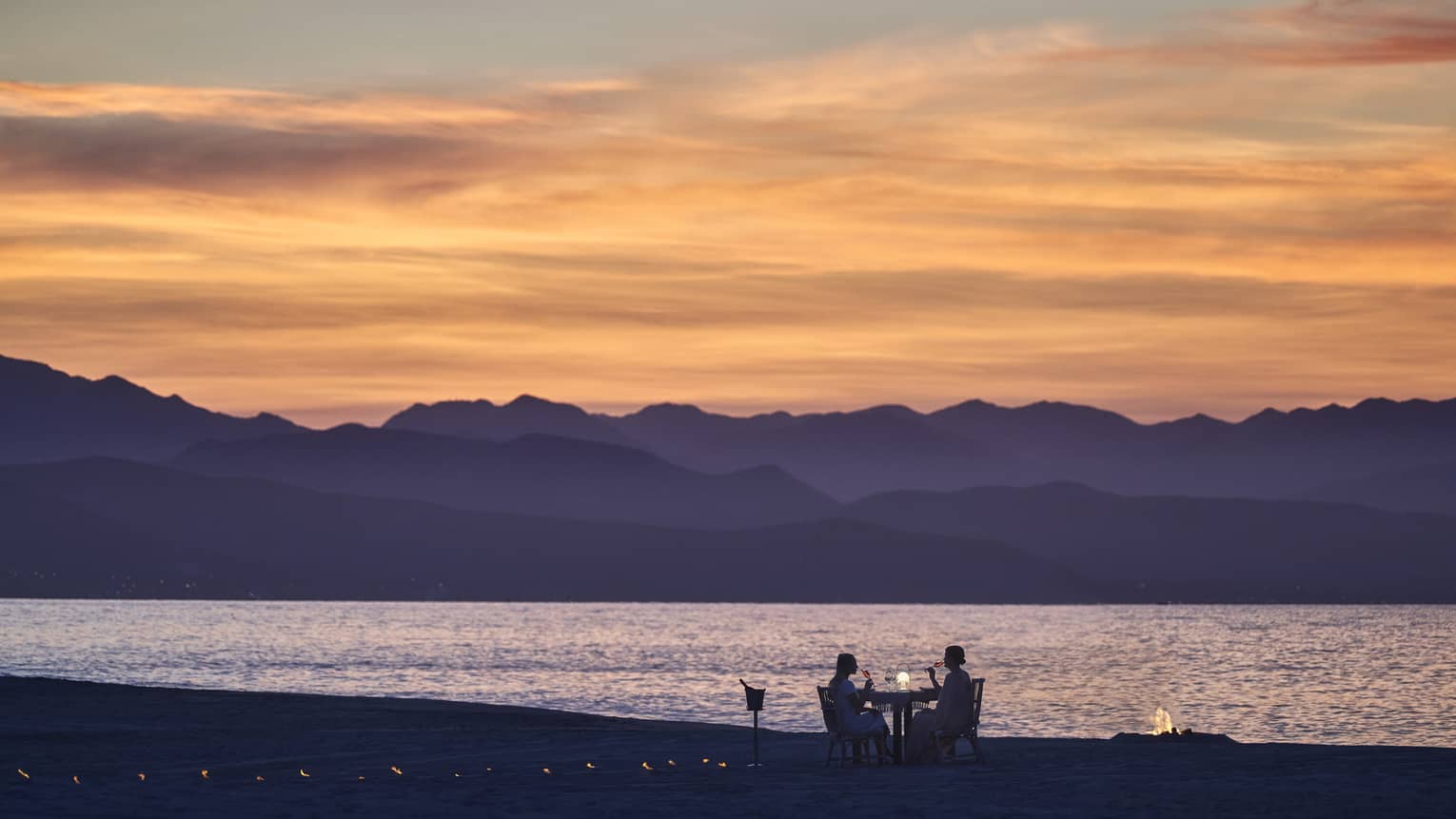 A couple dining on a beach during sunset, the mountains in the background look purple while the sky is orange and pink.