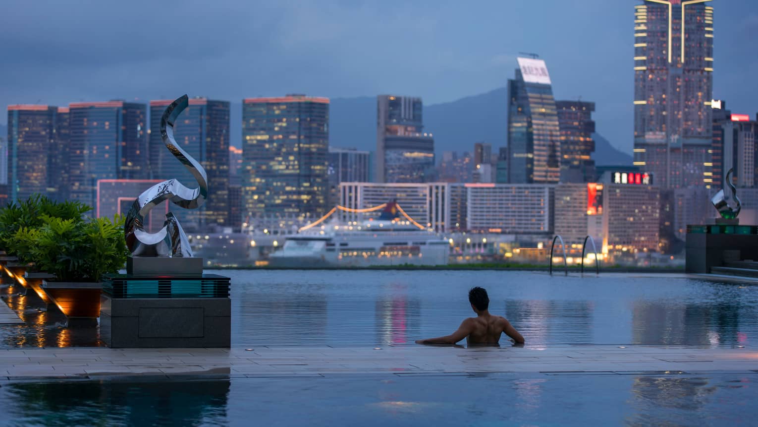 Silhouette of back of man wading in infinity swimming pool, looking out at Hong Kong harbour at dusk