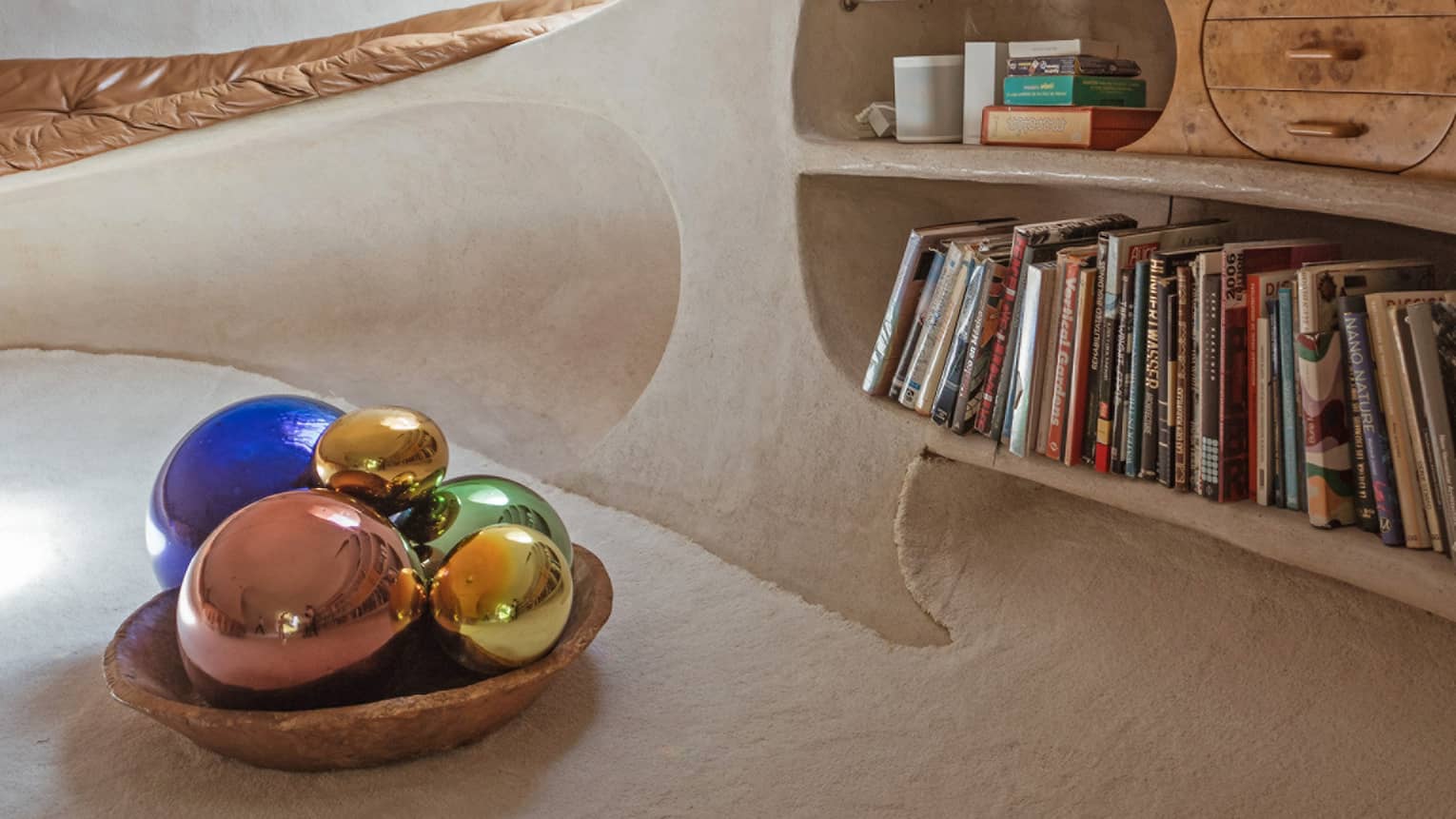 Aside a bowl of colourful orbs, shelves carved into a curved, adobe wall feature books, curios and inlaid wooden drawers. 
