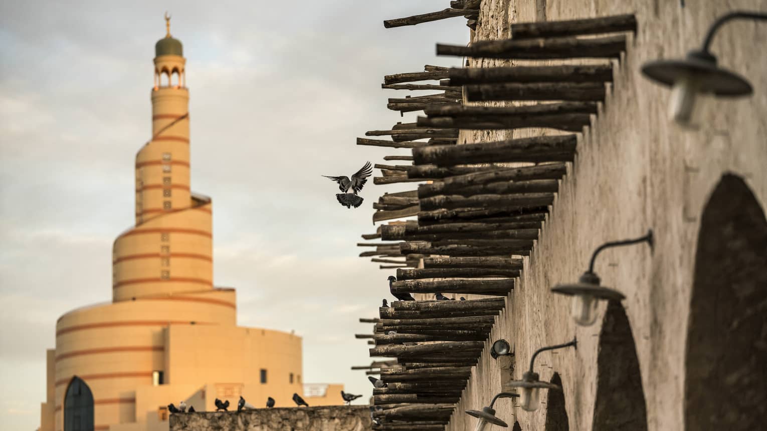 Old stone wall lined with narrow protruding logs, pigeons randomly flying and perched, a spiral minaret towering beyond.