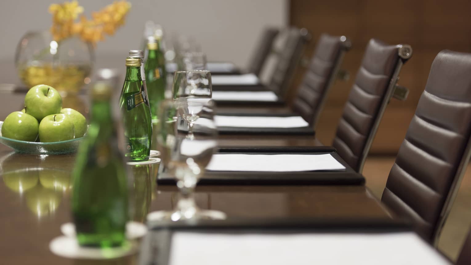 Close-up of business meeting table row of chairs, paper agendas, green glass bottles, apples