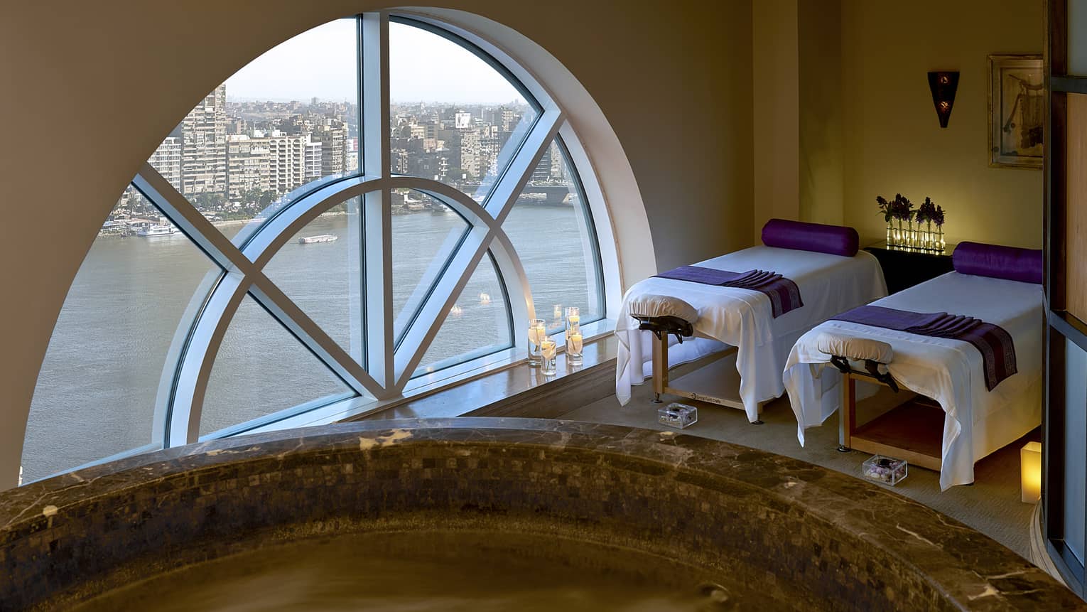 Two massage tables side-by-side in couples spa suite by lare crescent-shaped window overlooking Nile