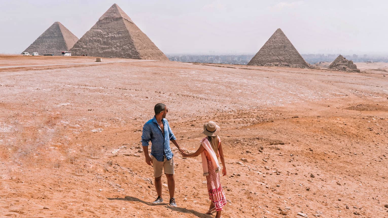 A couple holding hands turn back to look across the arid desert at the giant pyramids in the distance. 