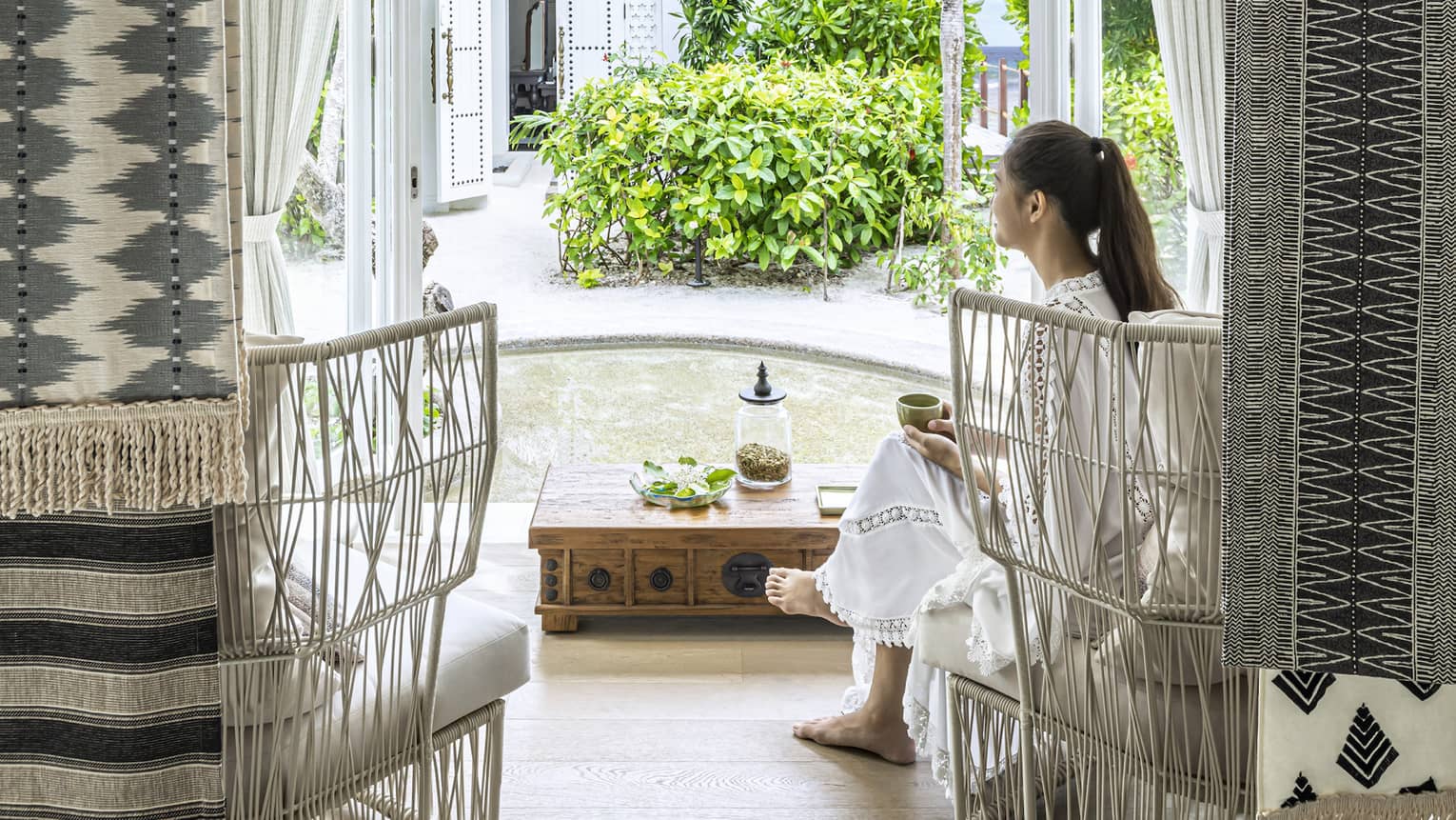 A woman sits in white wicker chair in the four seasons maldives spa waiting area and overlooks the garden outside an opened window