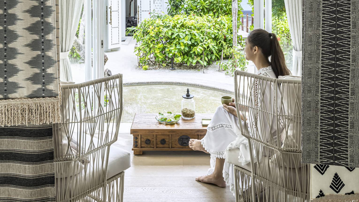 A woman sits in white wicker chair in the four seasons maldives spa waiting area and overlooks the garden outside an opened window