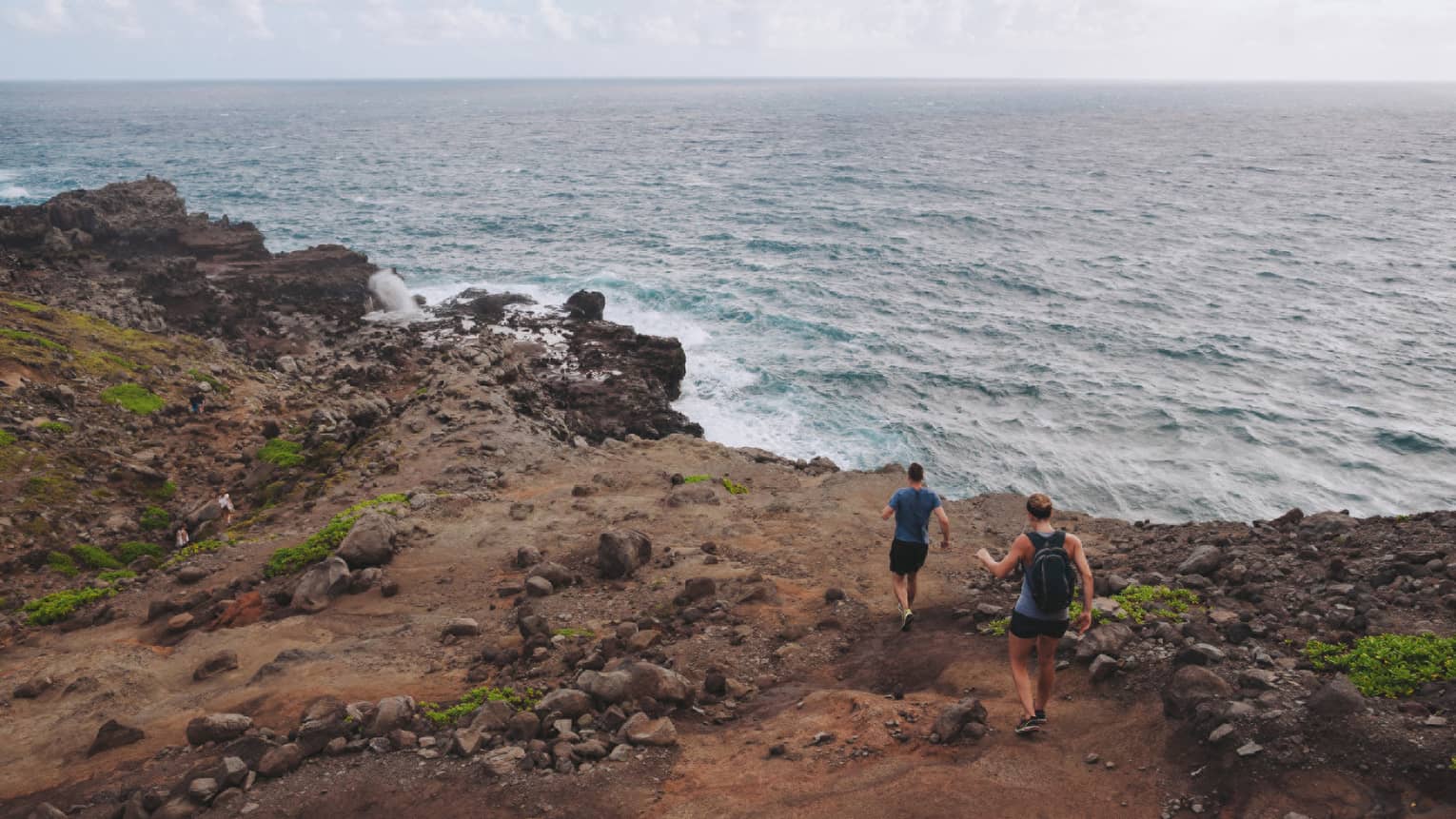 Two people hike down dirt-covered path, rocks towards ocean